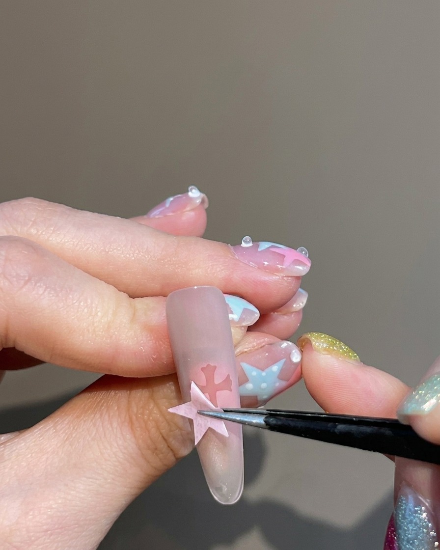 Eine Pinzette zieht einen Sticker von einem Nagel ab und hinterlässt eine klare, kreuzförmige Silhouette in der Mitte des blush-farbenen Nagels.