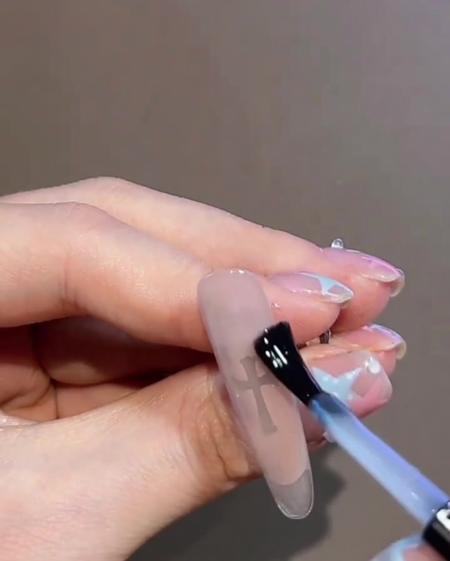 Eine Nahaufnahme eines klaren Gel-Top-Coats, der über ein fertiges Nageldesign mit Kreuz-Sticker und Strasssteinen aufgetragen wird.