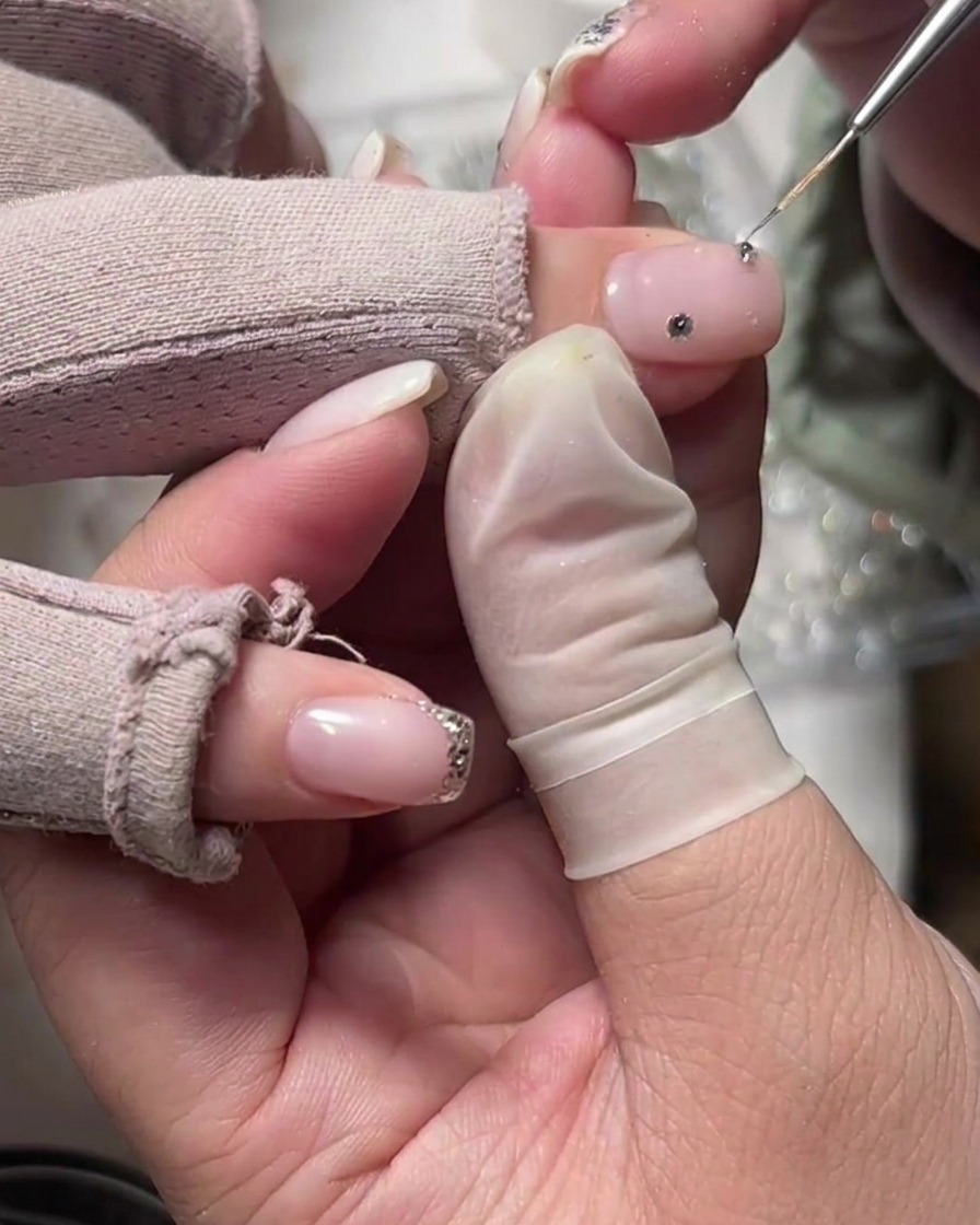 Ein feines Werkzeug platziert einen einzelnen Strassstein auf einem milchig-weißen Nagel in der Nähe des Nagelhautbereichs.