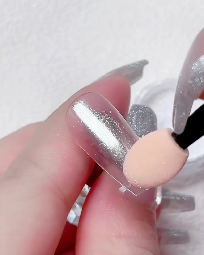 Eine Nahaufnahme eines Fingers, der einen transparenten Nageltip hält, während silbernes Chrom-Puder mit einem Lidschatten-Applikator aufgetragen wird.