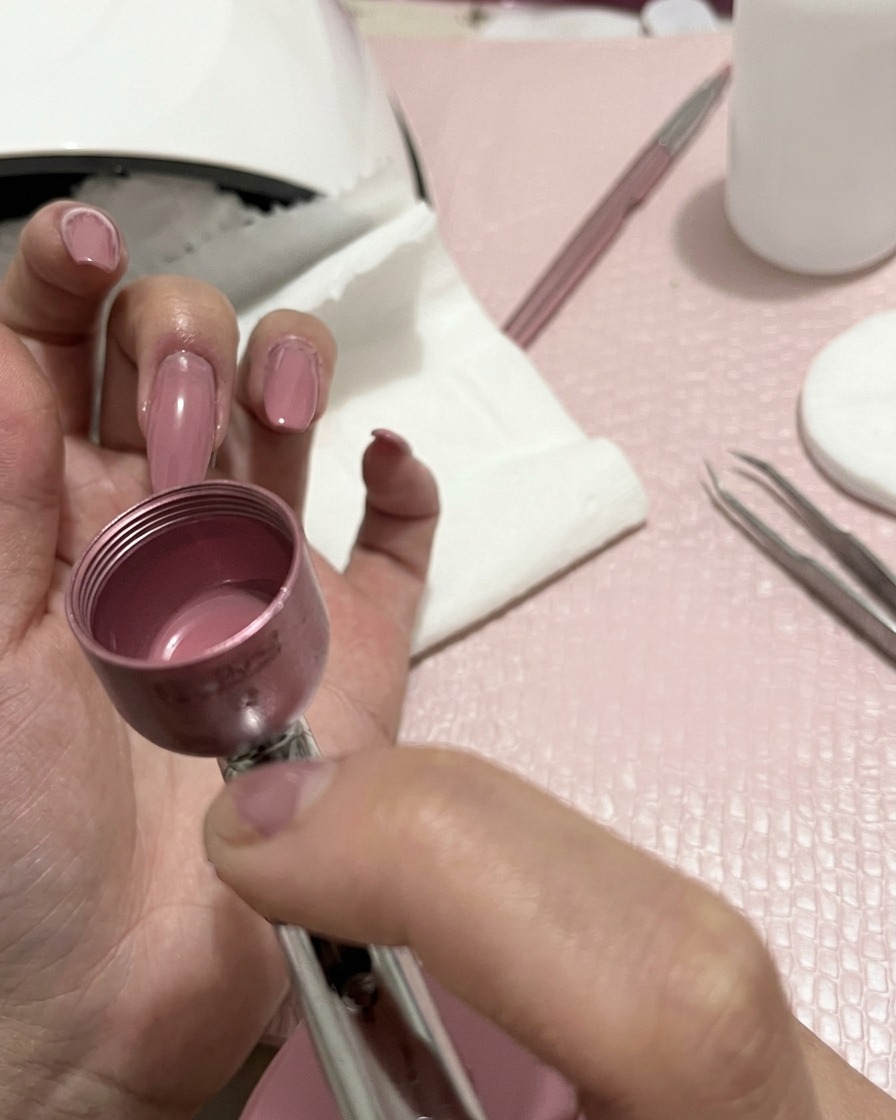 Eine Hand hält ein Airbrush-Gerät und sprüht einen feinen pfirsichfarbenen Nebel auf manikürte Fingernägel.