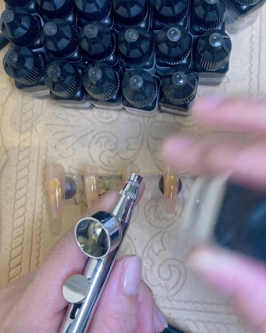 Eine Airbrush-Pistole, die einen sanften weißen Nebel auf die Spitze eines nude-farbenen Nagels aufträgt.