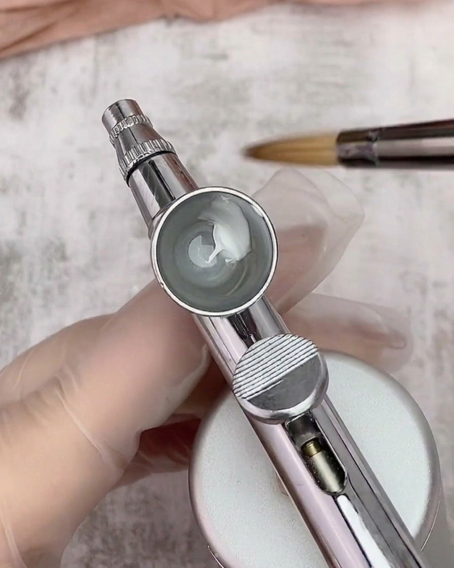 Blick in einen Airbrush-Becher mit einer Mischung aus weißem Gel und Verdünner, im Hintergrund ist ein Pinsel zu sehen.