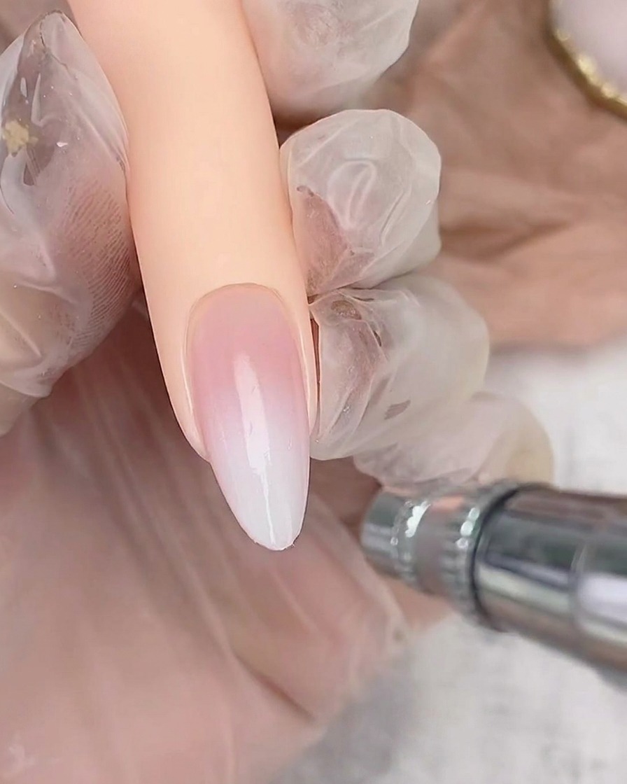 Ein Airbrush-Gerät sprüht leicht weiße Farbe auf die freie Kante eines mandelförmigen Nagels, um den Ombré-Verlauf aufzubauen.