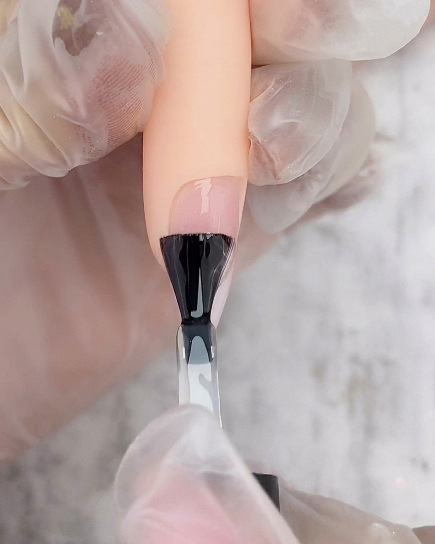 Ein schwarzer Nagellackpinsel trägt eine Schicht klaren Gel-Top-Coats über einem weiß-rosafarbenen Ombré-Nagel auf.