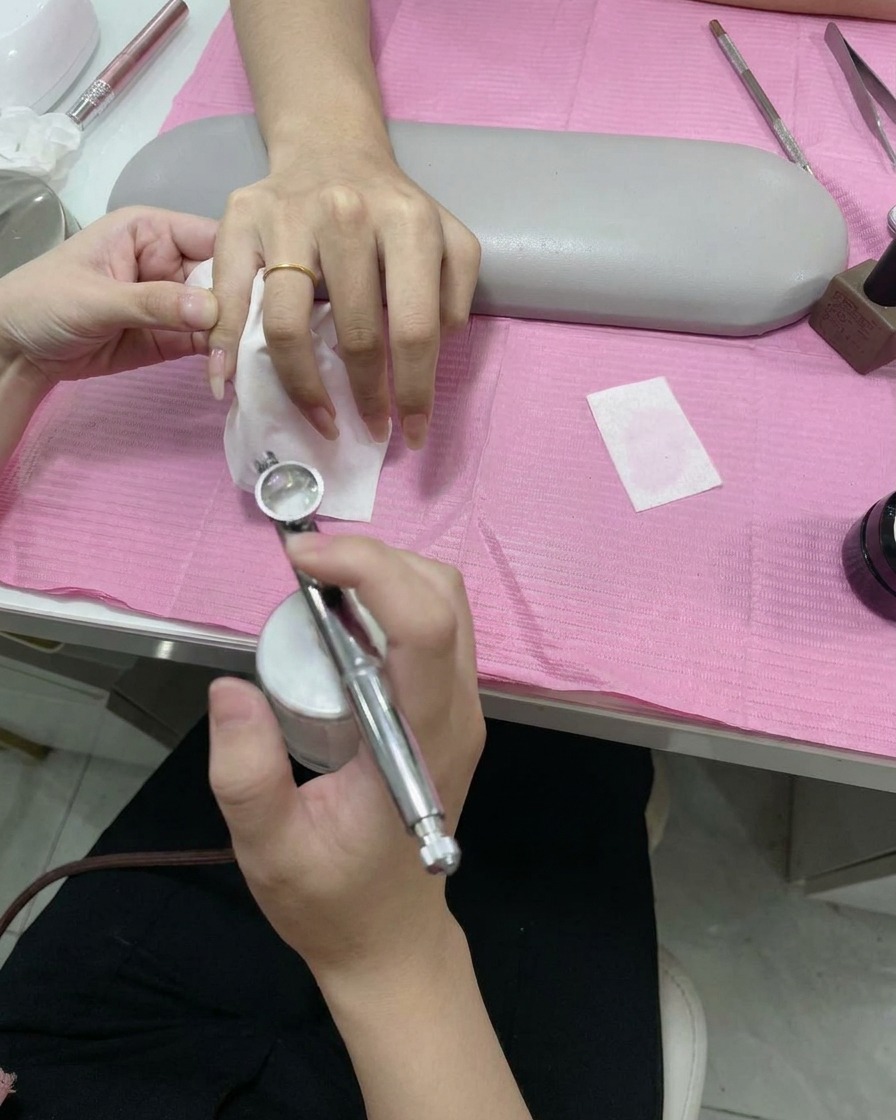 Eine Hand hält eine Airbrush-Pistole und sprüht einen feinen weißen Farbnebel über eine Schablone auf die Nagelspitze.