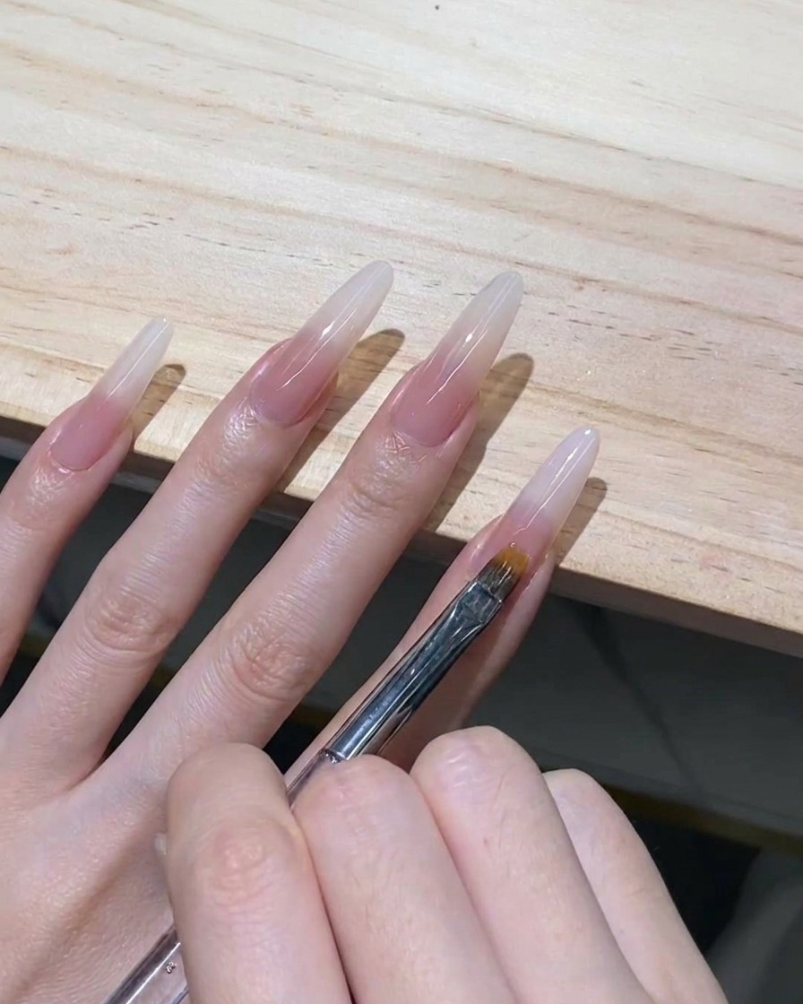Ein feinzahniger Ombré-Pinsel zieht sanft feuchtes, milchig-weißes Gel auf einer klaren Stiletto-Nagelverlängerung nach unten.