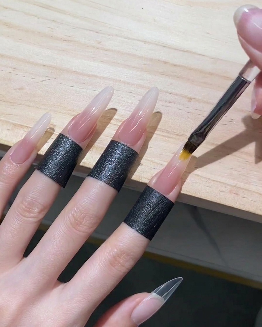 Ein Ombré-Pinsel verblendet zartes Nude-Gel nach oben in feuchtes weißes Gel auf einem Stiletto-Nagel, um einen nahtlosen Verlauf zu erzeugen.