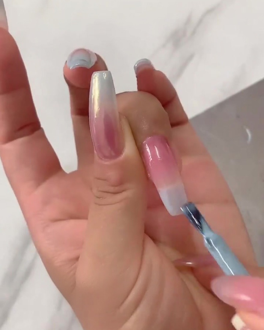 Nahaufnahme eines flachen Nagelpinsels, der konzentrierten hellblauen UV-Lack von der Spitze eines langen Fingernagels nach unten über eine zartrosa Basis verblendet.