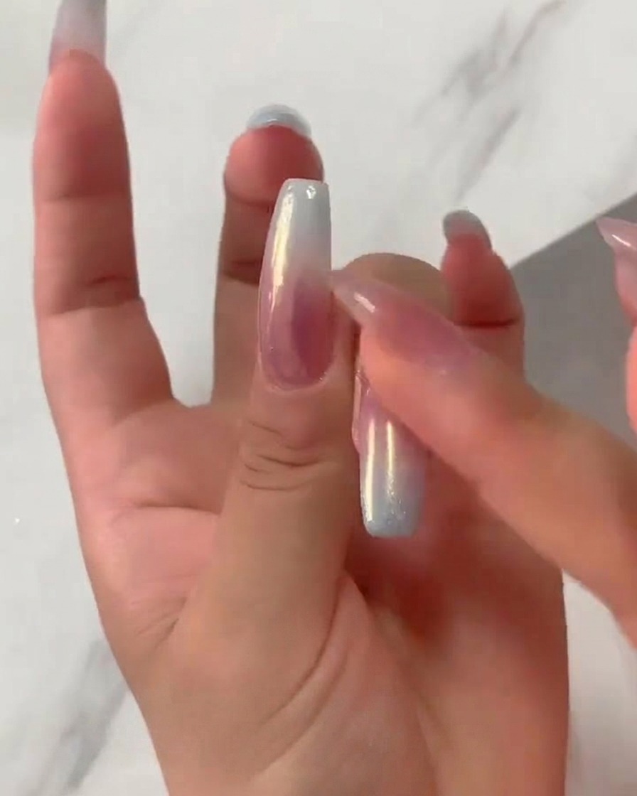 Eine Fingerspitze reibt fest irisierendes Aurora-Chromepuder über einen ausgehärteten Nagel mit rosa-blauem Farbverlauf.