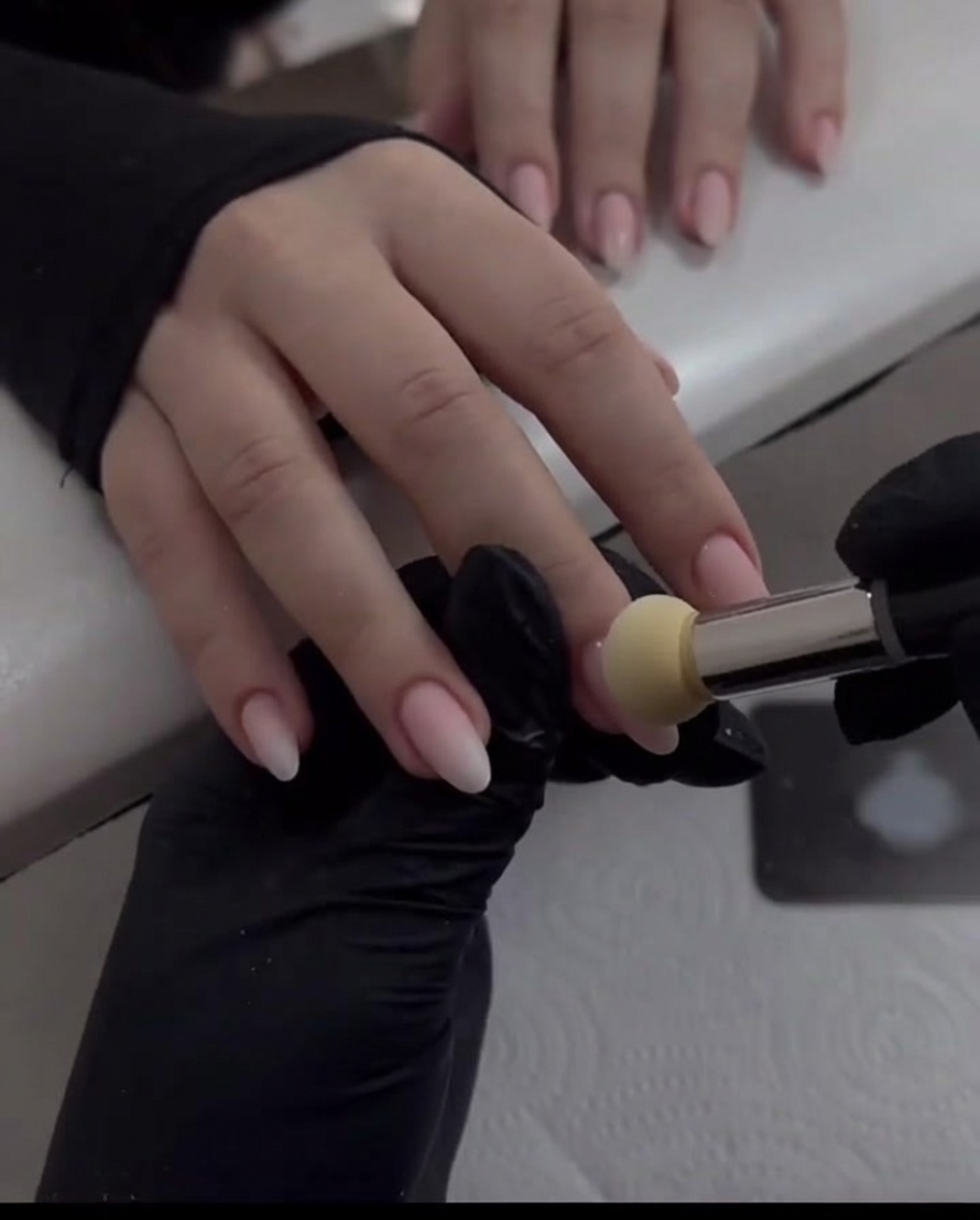 Eine behandschuhte Hand verwendet einen Schwammapplikator, um Pearl Chrome Puder auf einem nude-pinken Fingernagel nach unten zu verblenden.