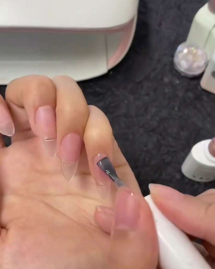 Eine Nahaufnahme von einem Pinsel, der ein halbdurchsichtiges Nude-Gel auf den Zeigefingernagel aufträgt.
