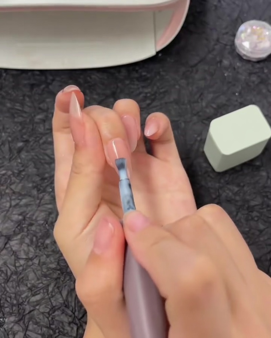 Ein Pinsel trägt einen horizontalen Streifen weißen Gel-Lacks auf die obere Hälfte eines Fingernagels auf.