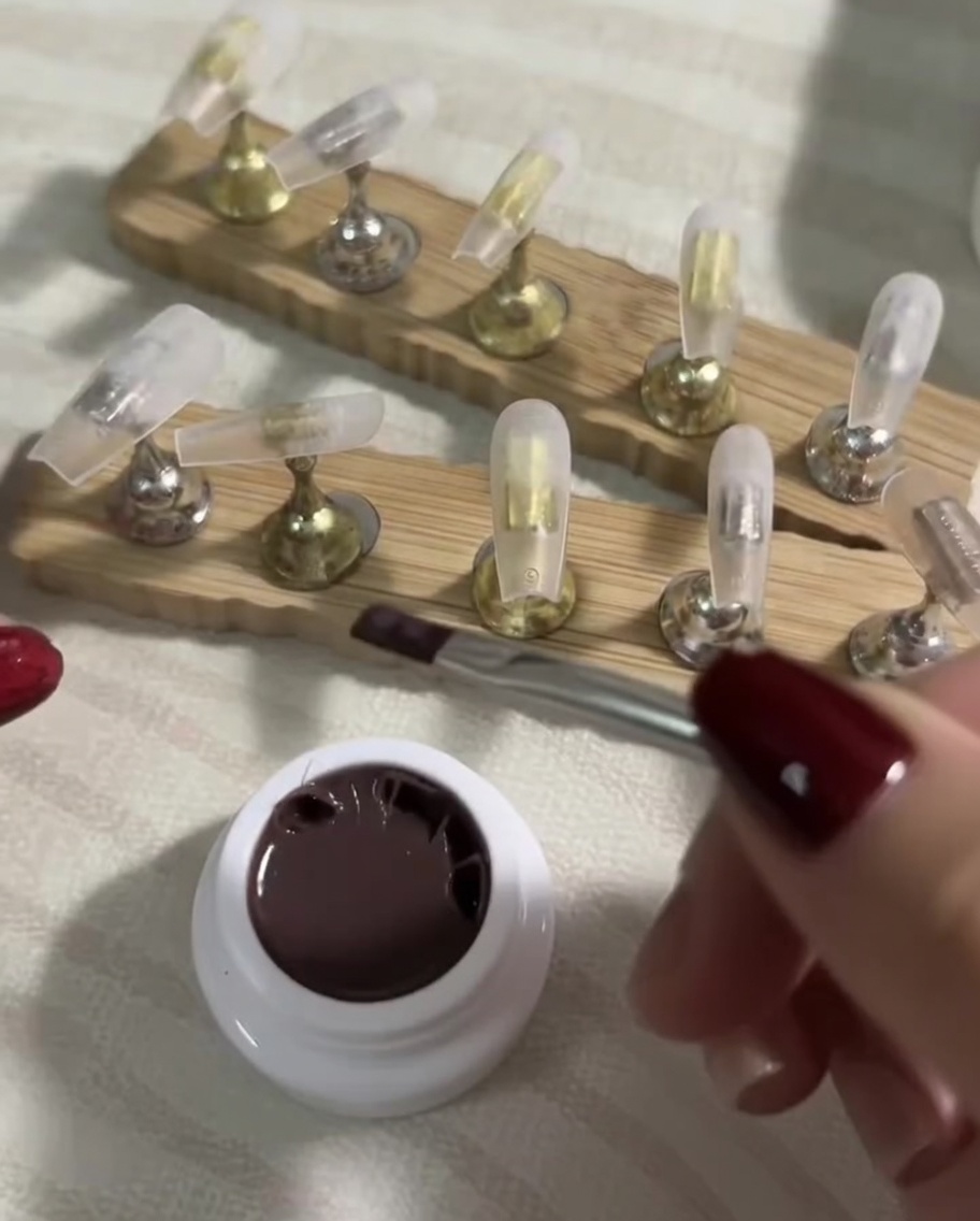 Ein flacher Pinsel trägt eine hauchdünne Schicht dunkles Nude-Gel auf einen klaren Press-On-Nageltipp auf.