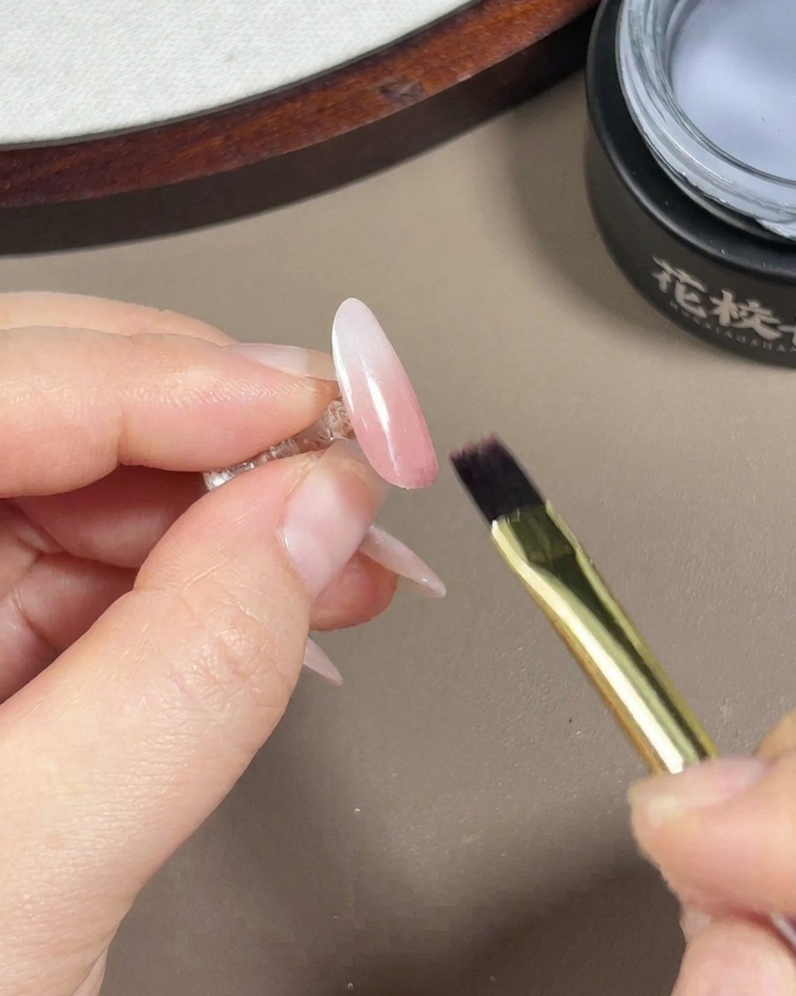 Ein Gel-Pinsel trägt einen klaren, glänzenden Top Coat auf einen fertigen Rosa-Weiß-Verlauf-Nagel auf.