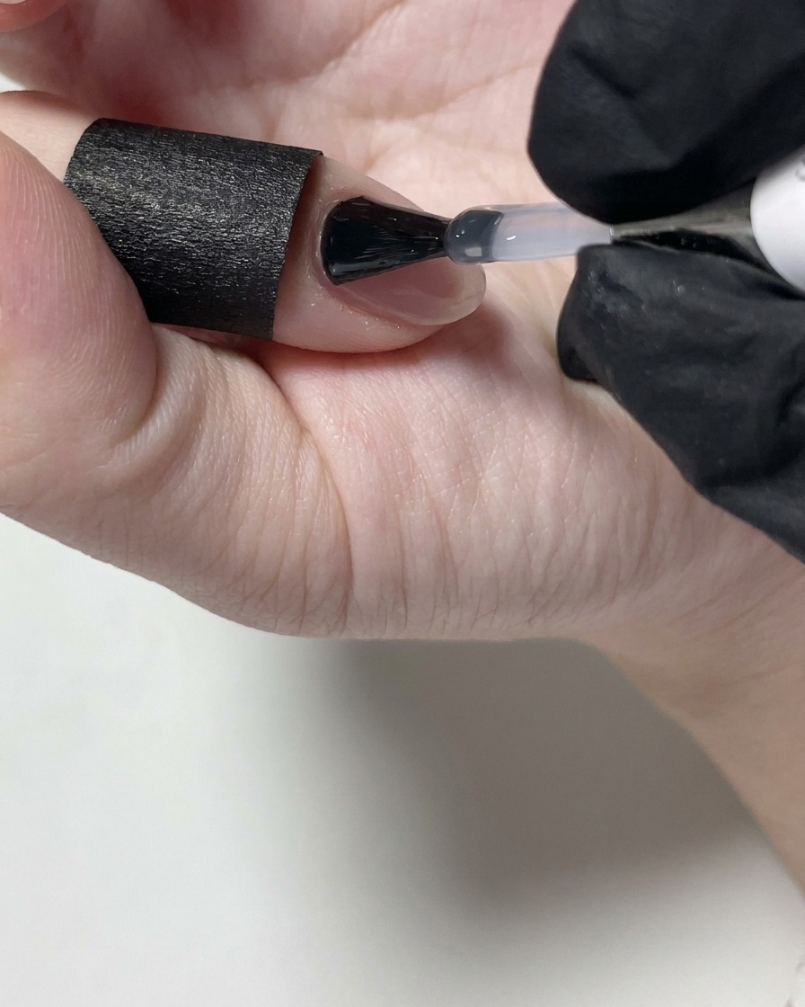 Ein Nageldesigner trägt mit einem schwarzen Pinsel matten Überlack auf einen nudefarbenen Gelnagel auf.
