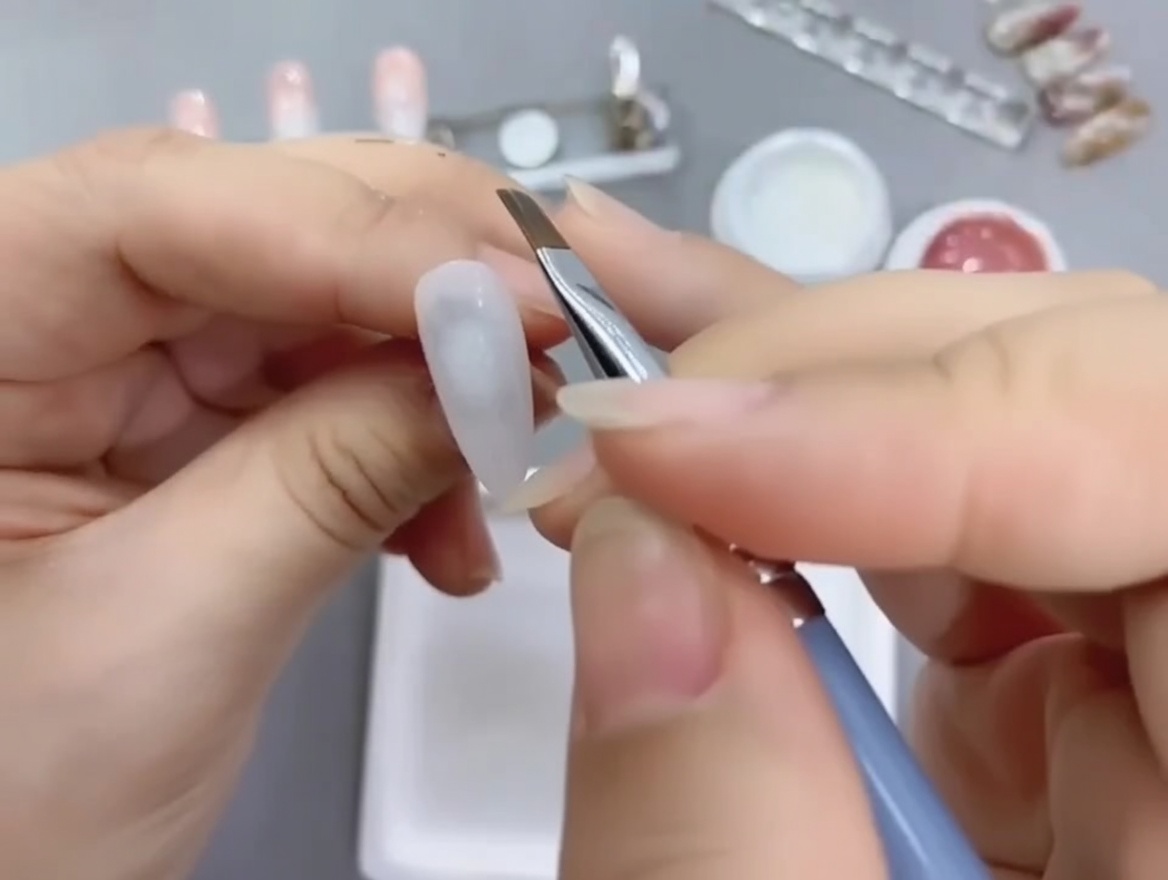 Ein abgewinkelter Nailart-Pinsel trägt Nude-Gel auf die untere Hälfte eines weiß lackierten Nageltips auf.