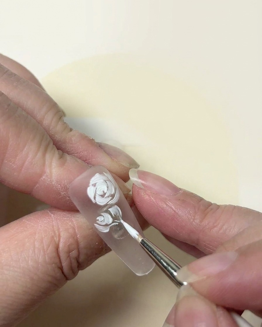 Nahaufnahme eines weißen Rosendesigns, das mit einem feinen Pinsel auf einem klaren Nagel verfeinert wird.