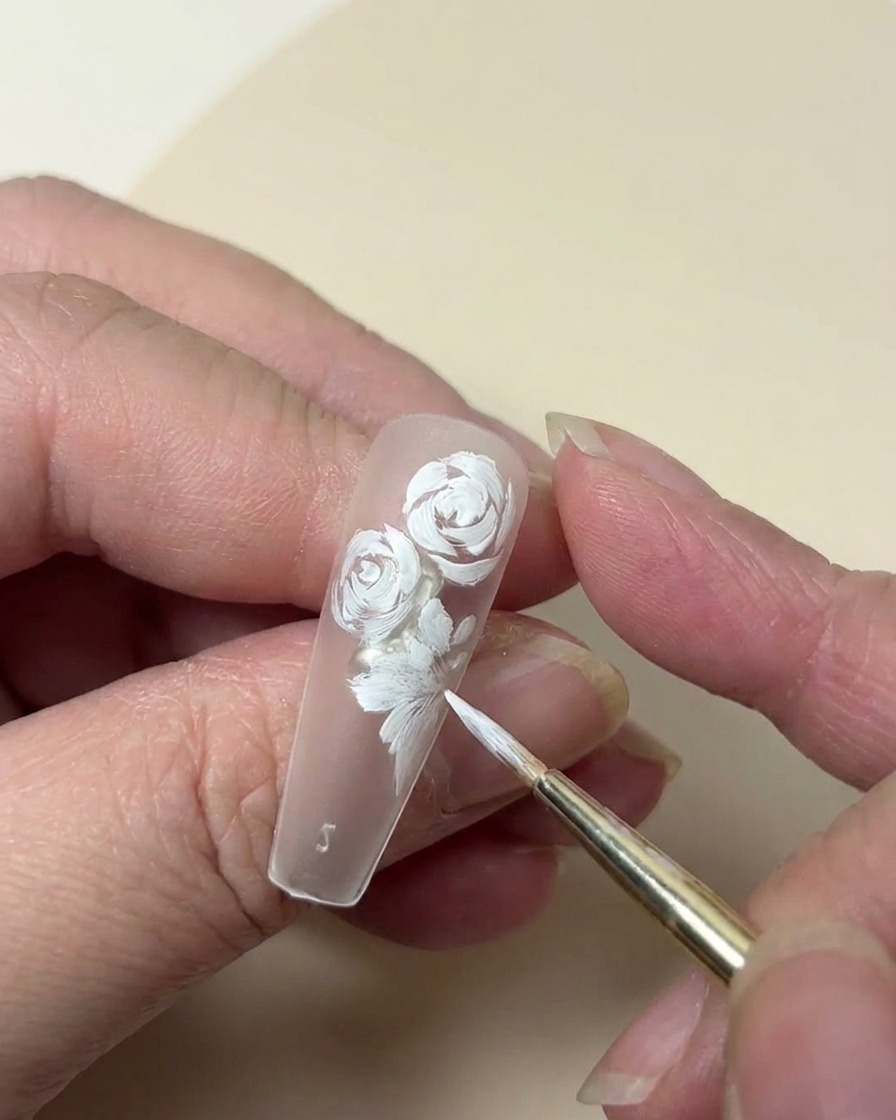 Nahaufnahme einer Nagelspitze, die eine weiße Rose ergänzt durch eine kleine fünfblättrige Blume und Blattdetails zeigt.