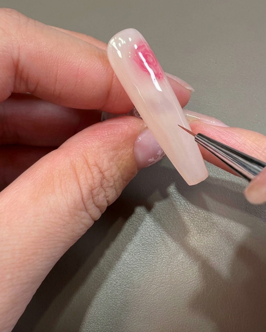 Ein feiner Pinsel arbeitet Details eines rosa, weichgezeichneten Bloom-Musters auf dem Nagel aus.