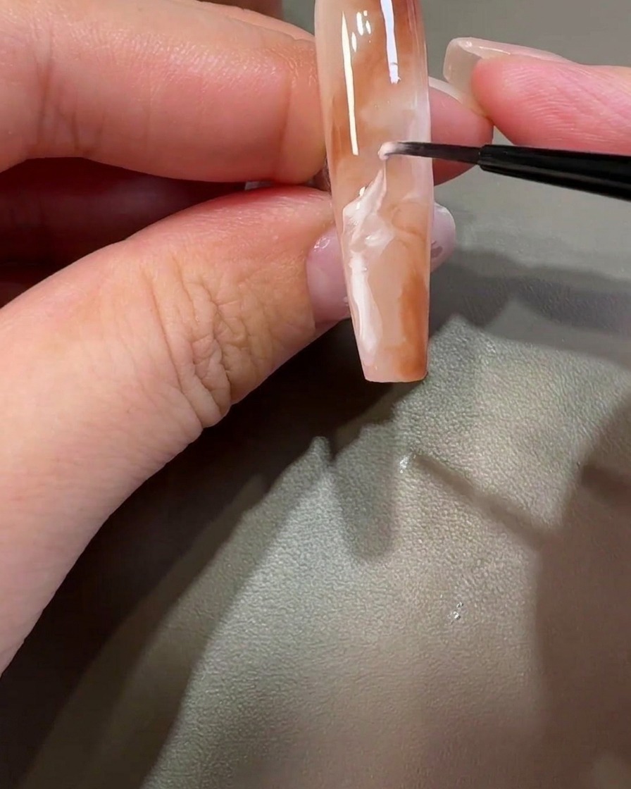 Ein feiner Pinsel zieht weißes Gel durch das braune Design, um wirbelnde Marmormuster zu erzeugen.