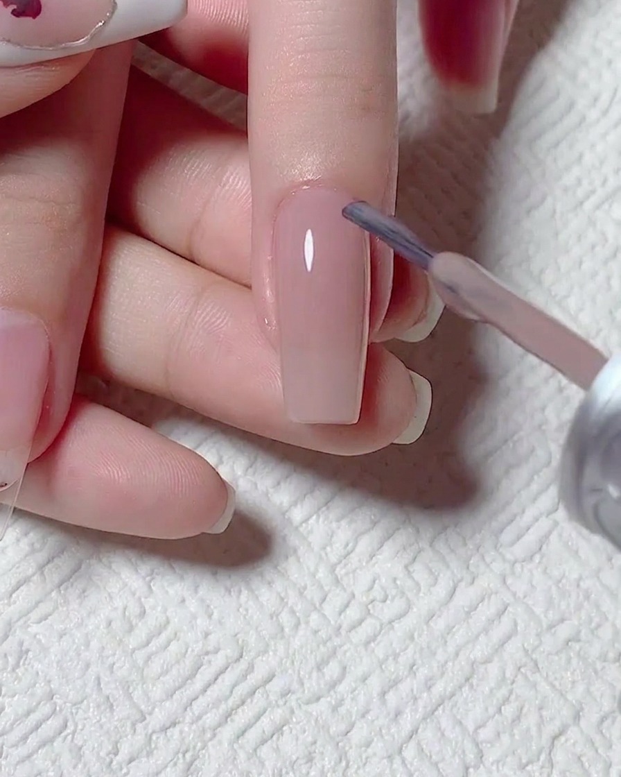 Nahaufnahme eines Pinsels, der ein transparentes, nudefarbenes Gel auf eine vorbereitete Nageloberfläche aufträgt.
