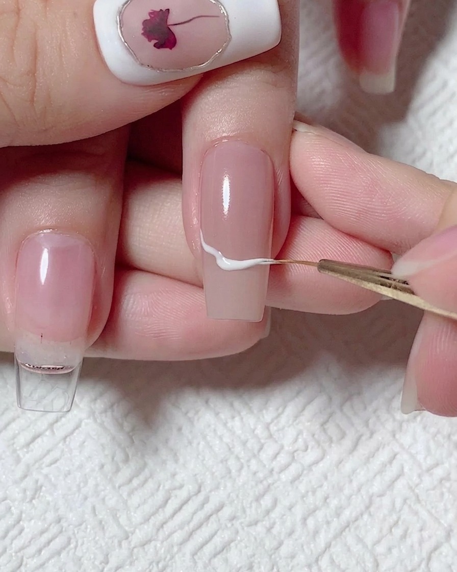 Ein feiner Liner-Pinsel zeichnet eine geschwungene weiße Linie quer über einen nudefarbenen Nagel.