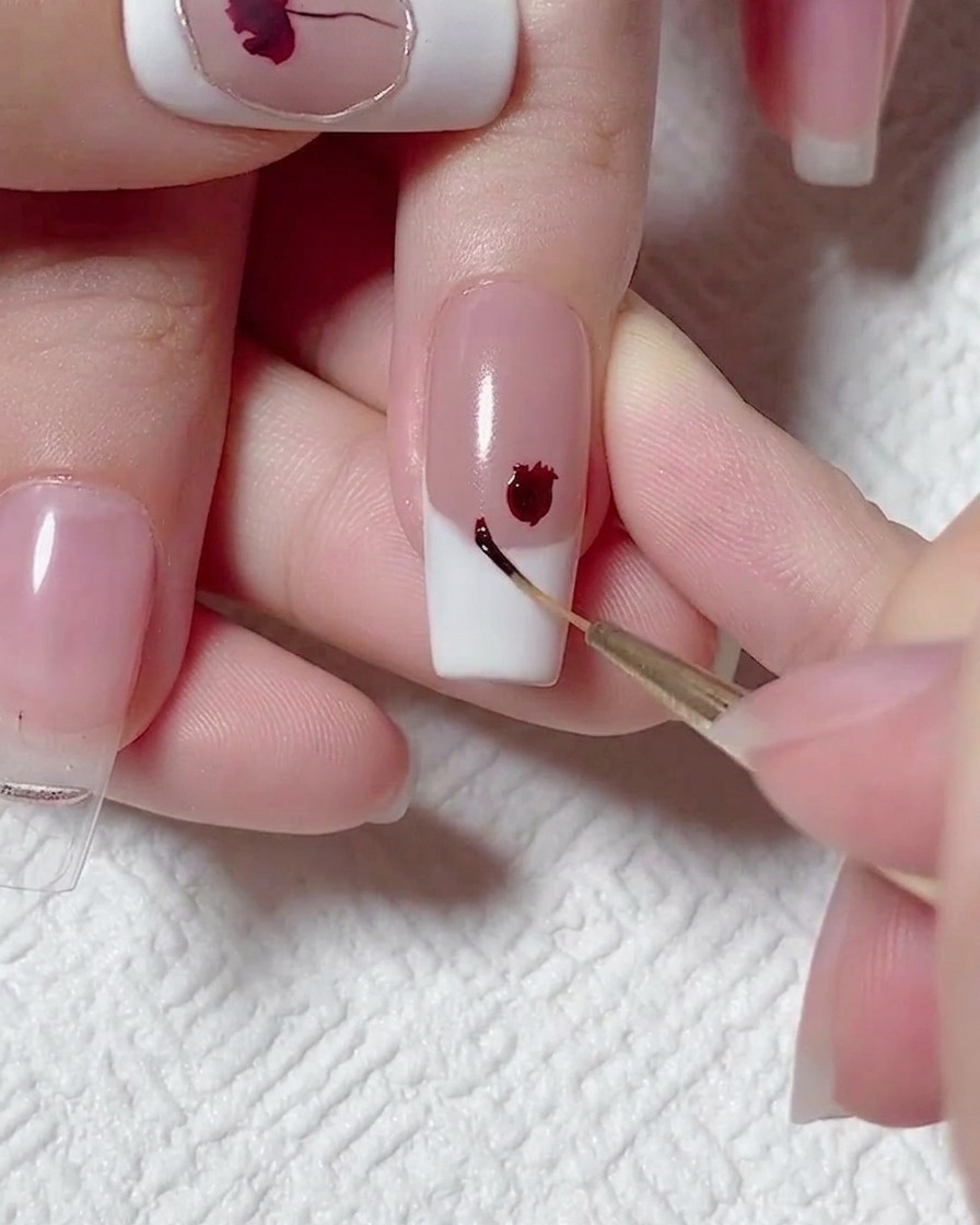 Nahaufnahme eines Liner-Pinsels, der einen winzigen Punkt aus dunkelrotem Gel neben einer größeren gemalten Blume auf einem French-Tip-Nagel anbringt.