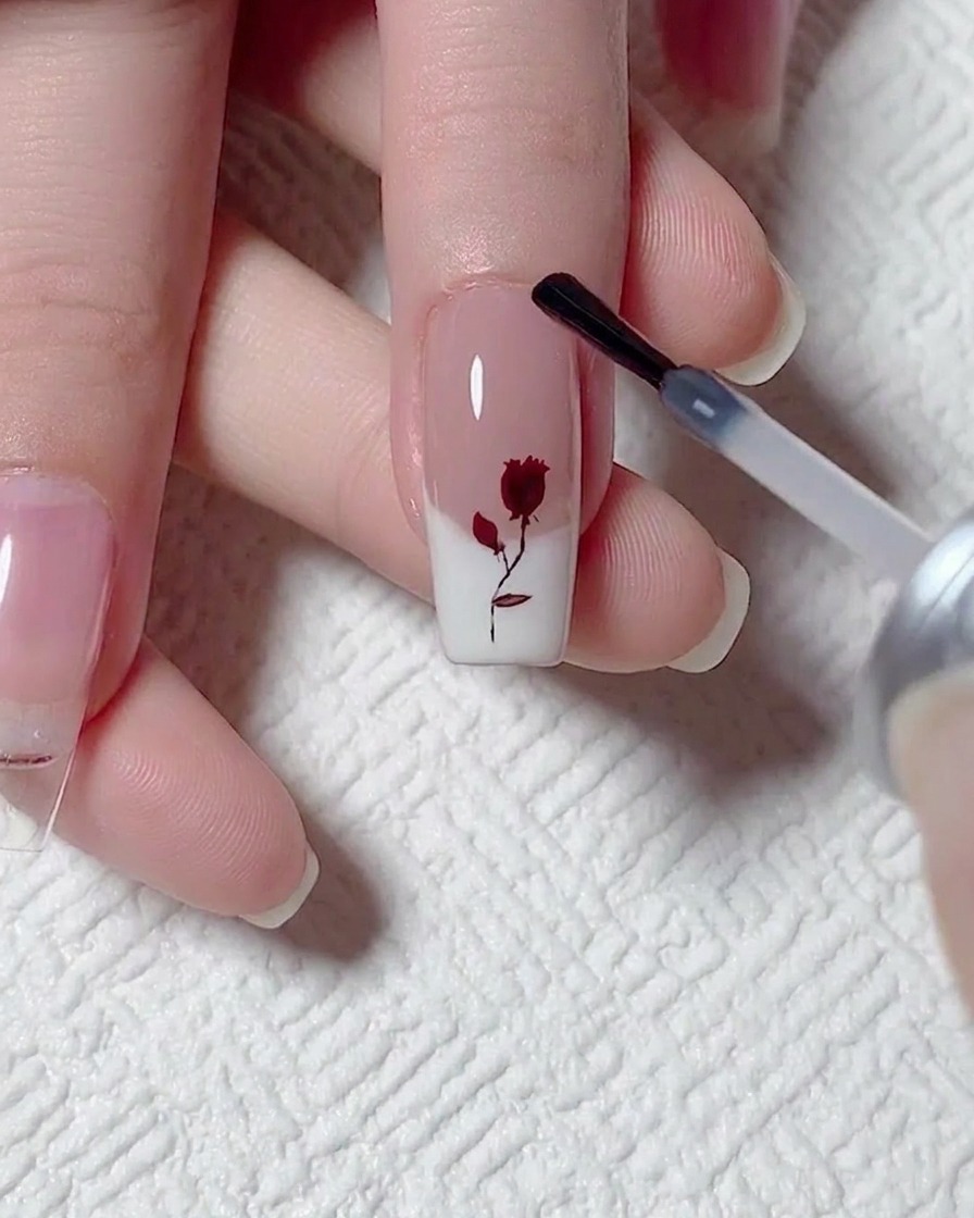 Auftragen einer Schicht mattem Top Coat mit einem Pinsel über einen Nagel mit rotem Rosen- und weißem French-Design.