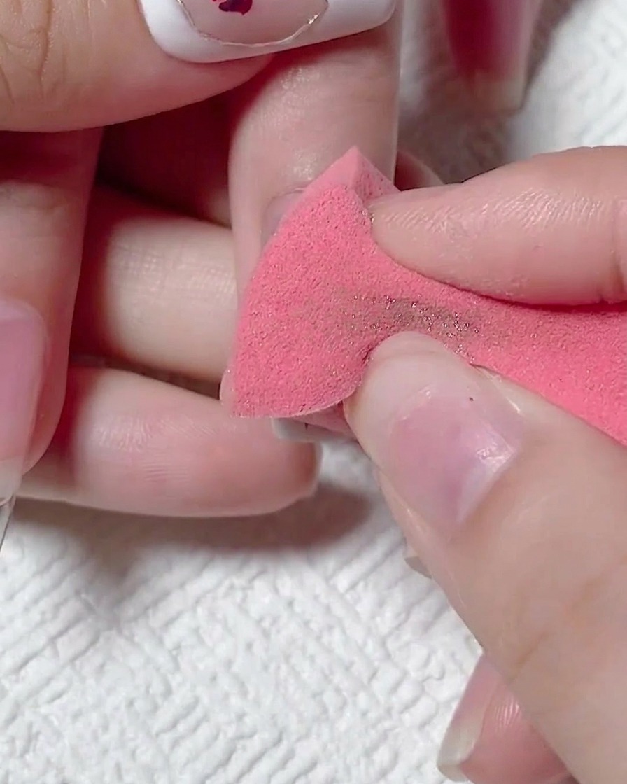 Eine Hand benutzt einen trockenen rosa Make-up-Schwamm, um überschüssiges goldenes Chrompuder von einem matten Nagel abzuwischen.