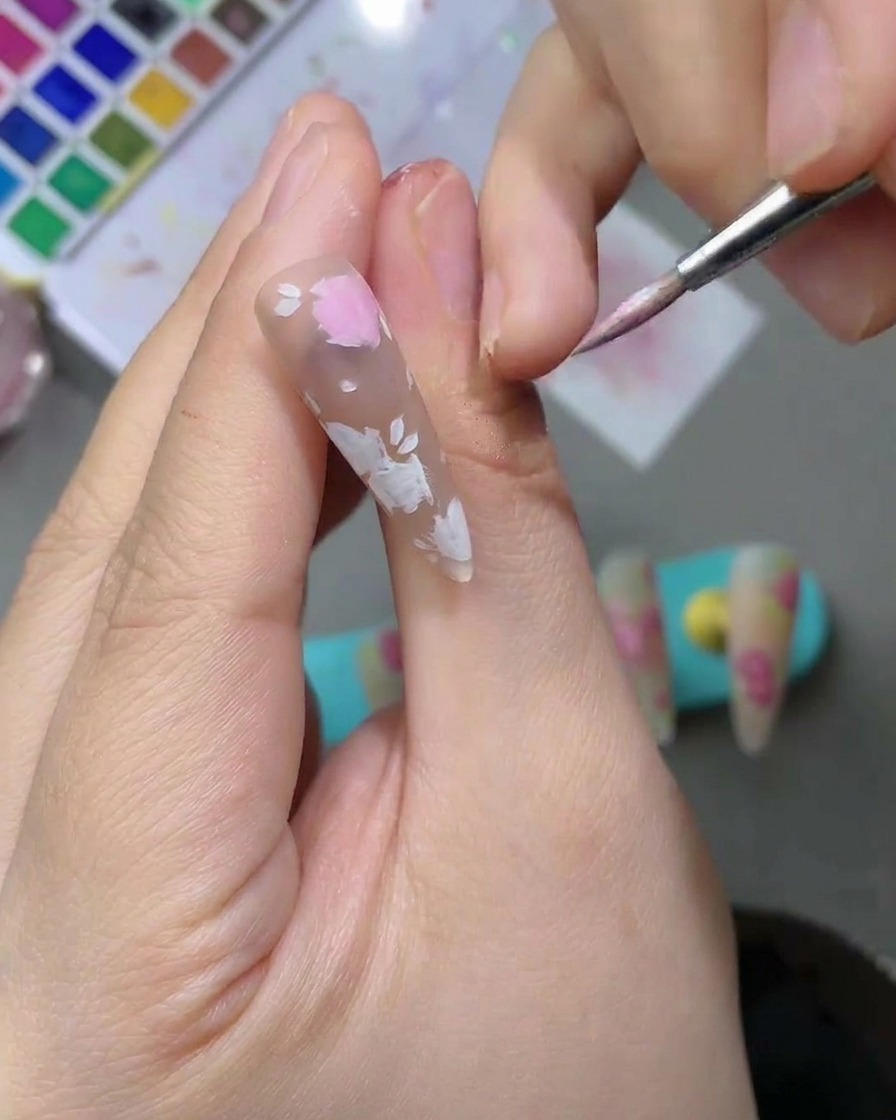 Nahaufnahme eines Nagels, bei dem zartrosa Aquarell-Blütenformen mit einem feinen Pinsel ausgefüllt werden.