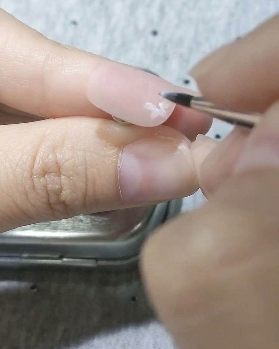 Nahaufnahme eines feinen Detailpinsels, der halbtransparente weiße Blütenblätter auf eine matte rosa Nagelbasis malt.