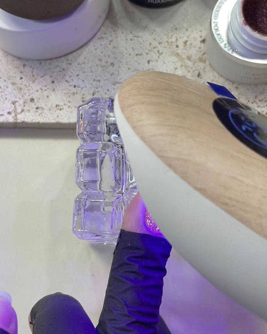 Eine Hand wird unter einer UV/LED-Lampe platziert, während ein Nagel mit rosa Glitzer aushärtet.
