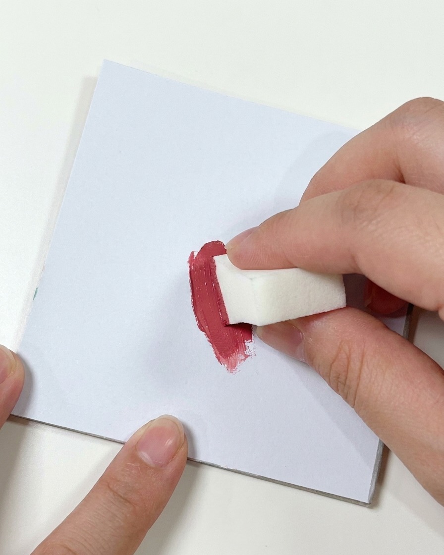 Eine Hand drückt einen kleinen weißen Make-up-Schwamm in einen Klecks dunkles Pink-Gel auf einer flachen weißen Oberfläche.