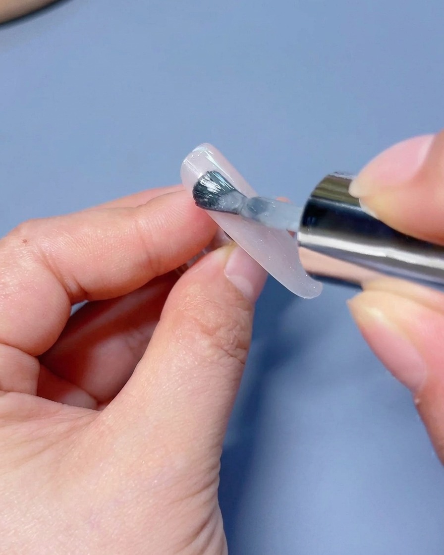 Ein Pinsel trägt schimmerndes, weißes Cat-Eye-Gel auf einen Press-on-Nagel auf.