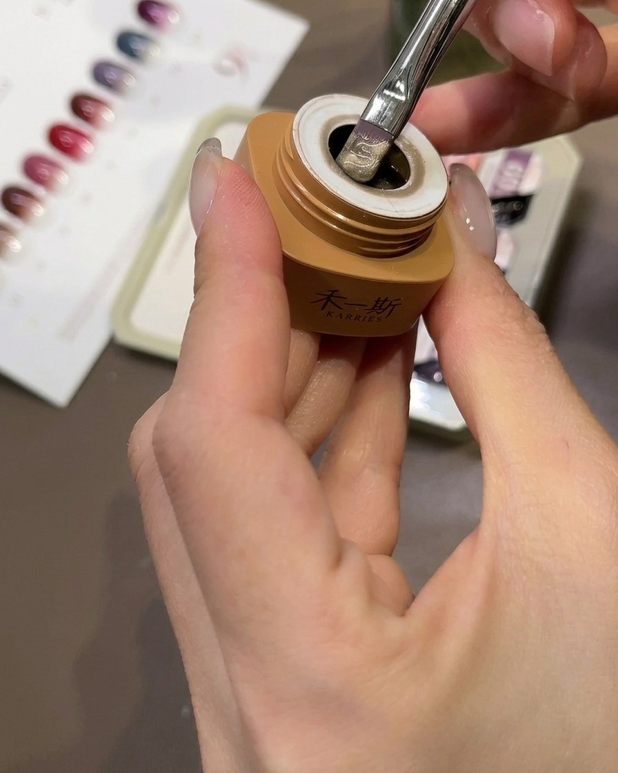 Ein Pinsel trägt beigefarbenes Katzenaugen-Gel auf einen klaren Nagel-Tip auf.