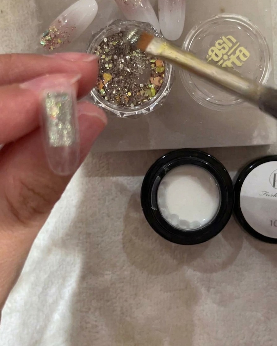 Ein Pinsel trägt groben silbernen und irisierenden Glitzer auf die obere Hälfte eines transparenten Nageltips auf.