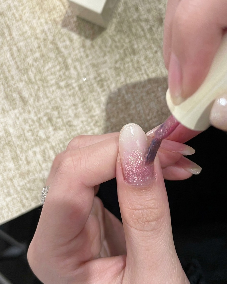 Ein Pinsel trägt ein zweites, etwas stärker pigmentiertes rosa Glitzergel auf den Nagel auf.