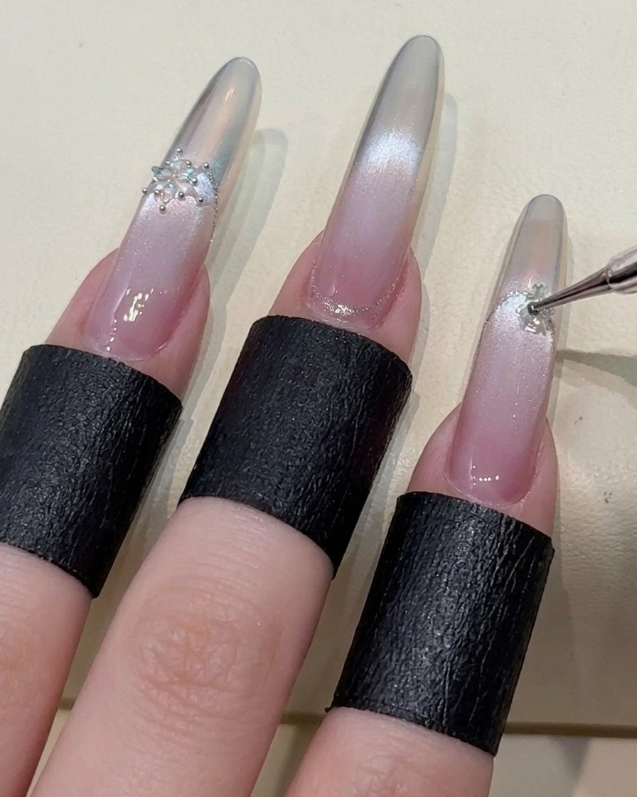 Ein Zeigefinger wird mit einem Schneeflocken-Kristall verziert, was den detaillierten Nail-Art-Prozess unterstreicht.