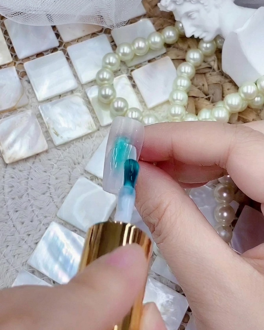 Ein Pinsel trägt eine Schicht milchig-weißes Gel-Polish auf einen transparenten Press-on-Nageltipp auf.