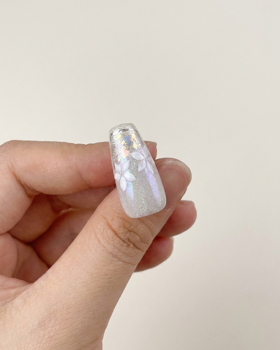 Ein Pinsel trägt eine Schicht klaren, hochglänzenden Top Coat über ein fertiges Nageldesign mit weißen Blumen und Silber-Flocken auf.