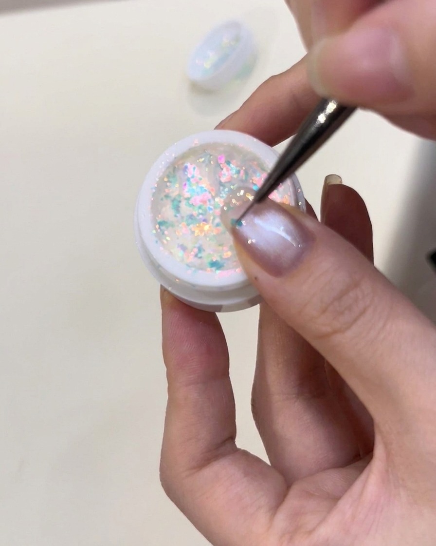 Eine Pinzette platziert vorsichtig kleine, blau leuchtende Flakies auf einem mit Aufbaugel beschichteten Nagel.
