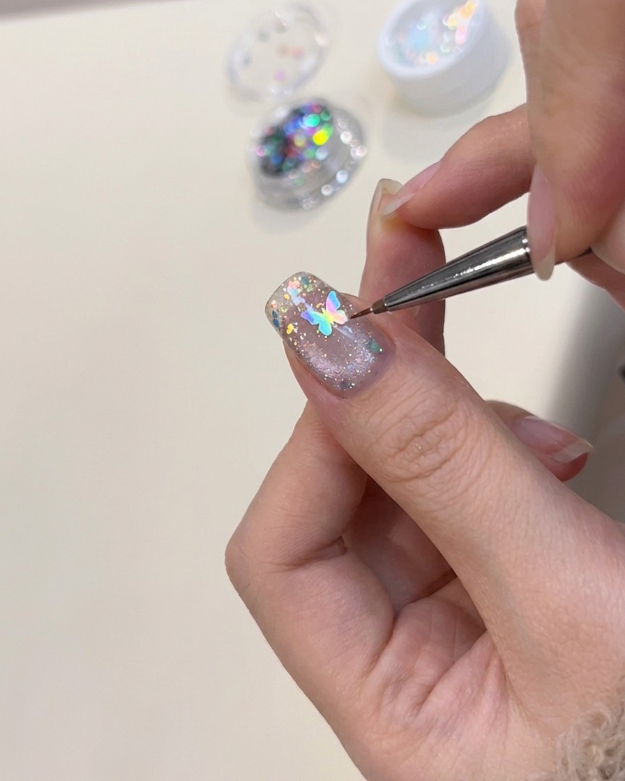 Eine Pinzette positioniert einen kleinen, holografischen Schmetterlings-Charm auf einem dekorierten Nagel.