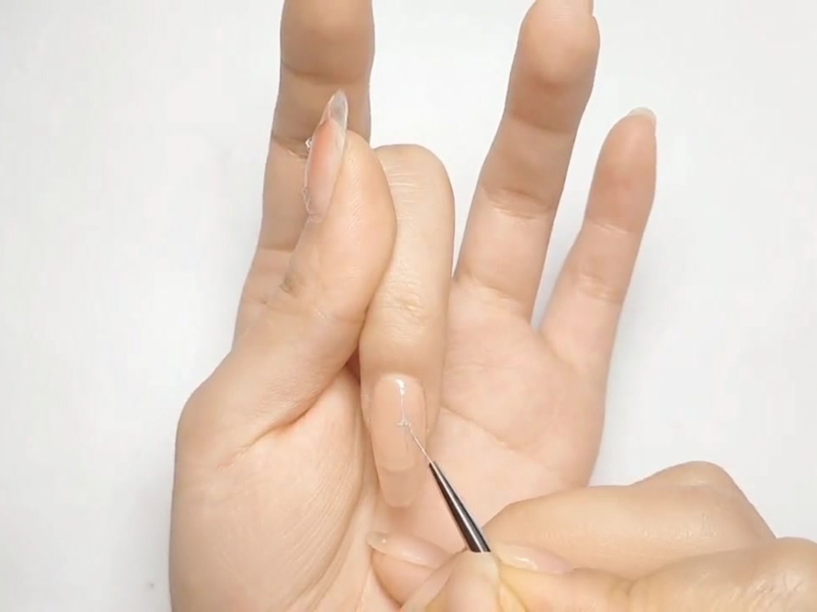 Ein dünner Nailart-Pinsel zeichnet ein silbernes Kreuz auf einen nudefarbenen Nagel.