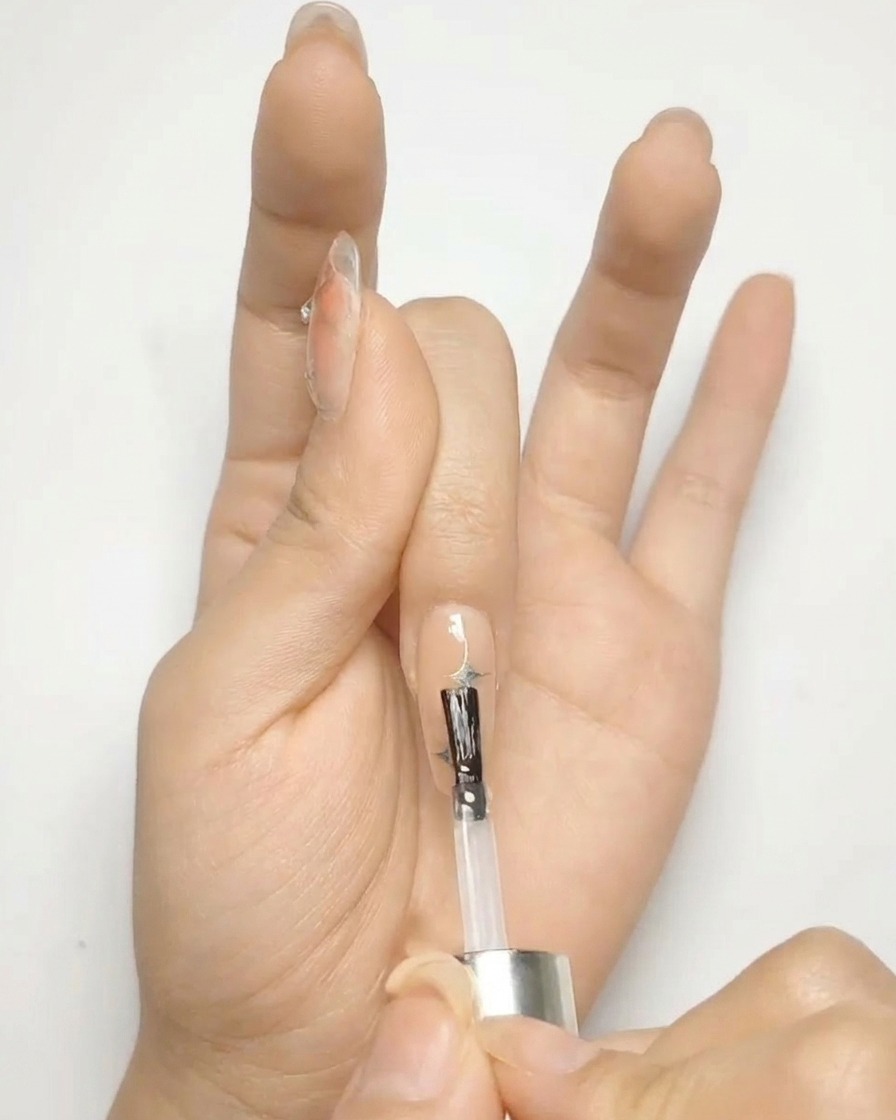 Ein Pinsel trägt klaren Hochglanz-Top-Coat über einen Nude-Nagel mit silbernen Sternen auf.
