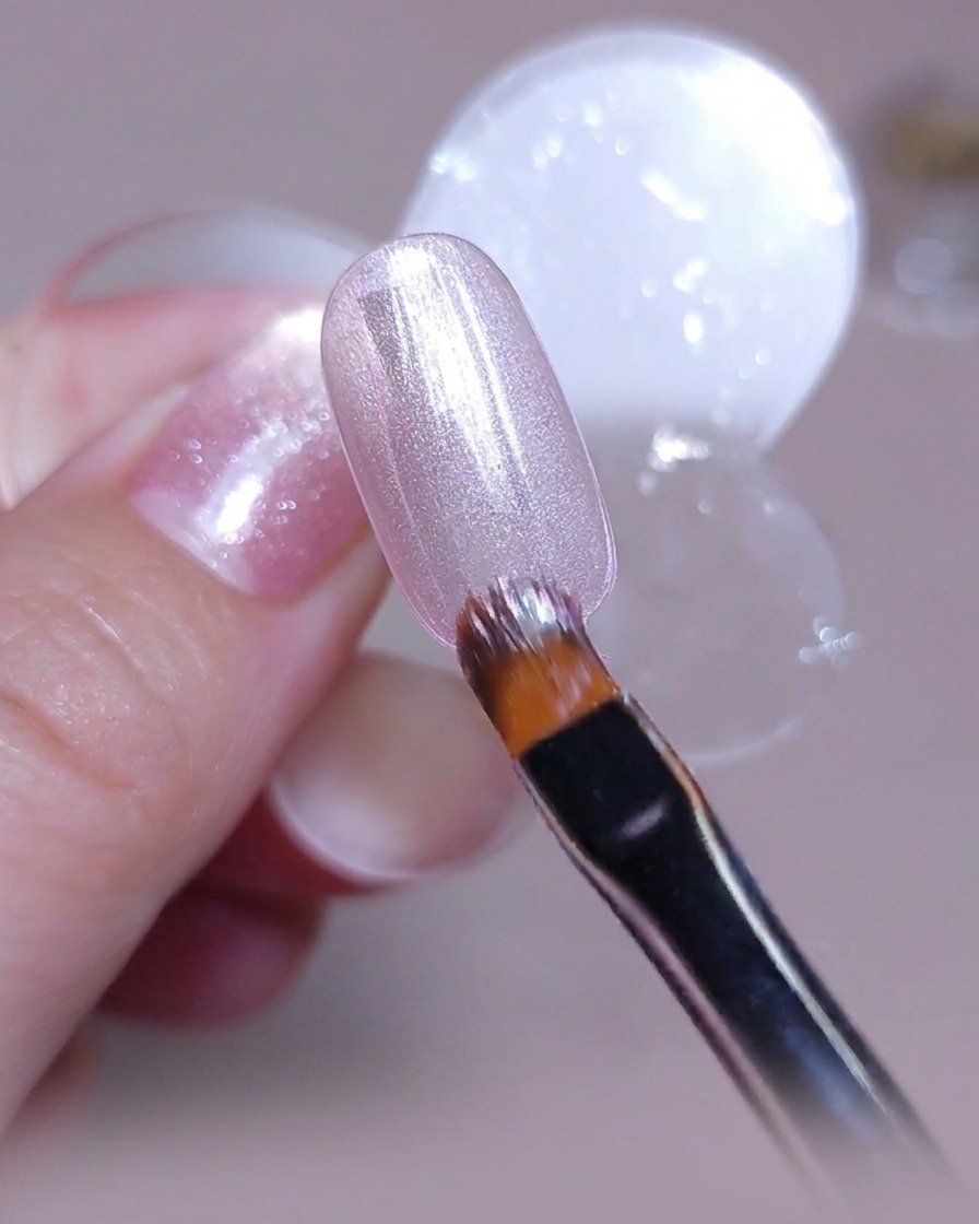 Ein Pinsel trägt eine gleichmäßige Schicht pink-silbernen Cat-Eye-Gellacks auf einen klaren Nagel-Tip auf.
