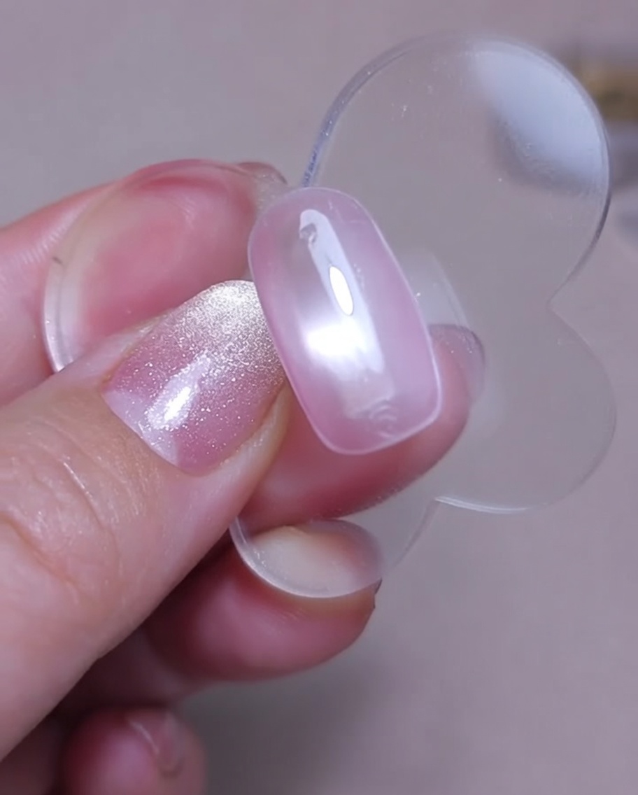 Nahaufnahme eines magnetischen Nagelwerkzeugs, das über einen feuchten pinken Cat-Eye-Nagel gehalten wird, um einen Schimmereffekt zu erzielen.