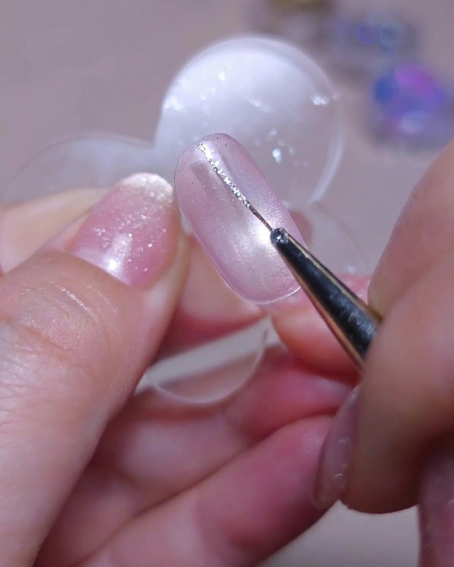 Ein feiner Detailpinsel zeichnet eine zarte vertikale Linie mit Silber-Glitzergel auf der linken Seite eines Cat-Eye-Nagels.