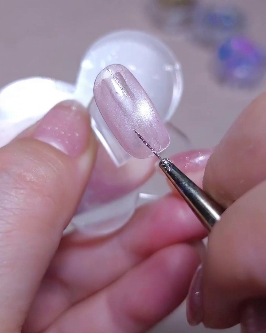 Ein feiner Liner-Pinsel zeichnet eine zweite gerade vertikale Silber-Glitzerlinie neben eine bestehende Linie auf einem schimmernden pinken Cat-Eye-Nagel.
