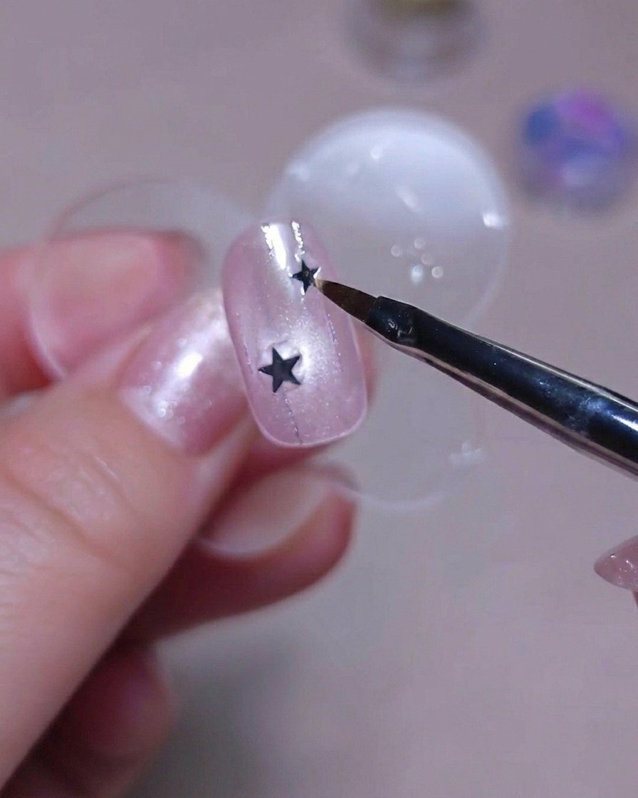 Nahaufnahme einer Pinselspitze, die eine winzige silberne Stern-Paillette über einem pinken glitzernden Nagel hält.